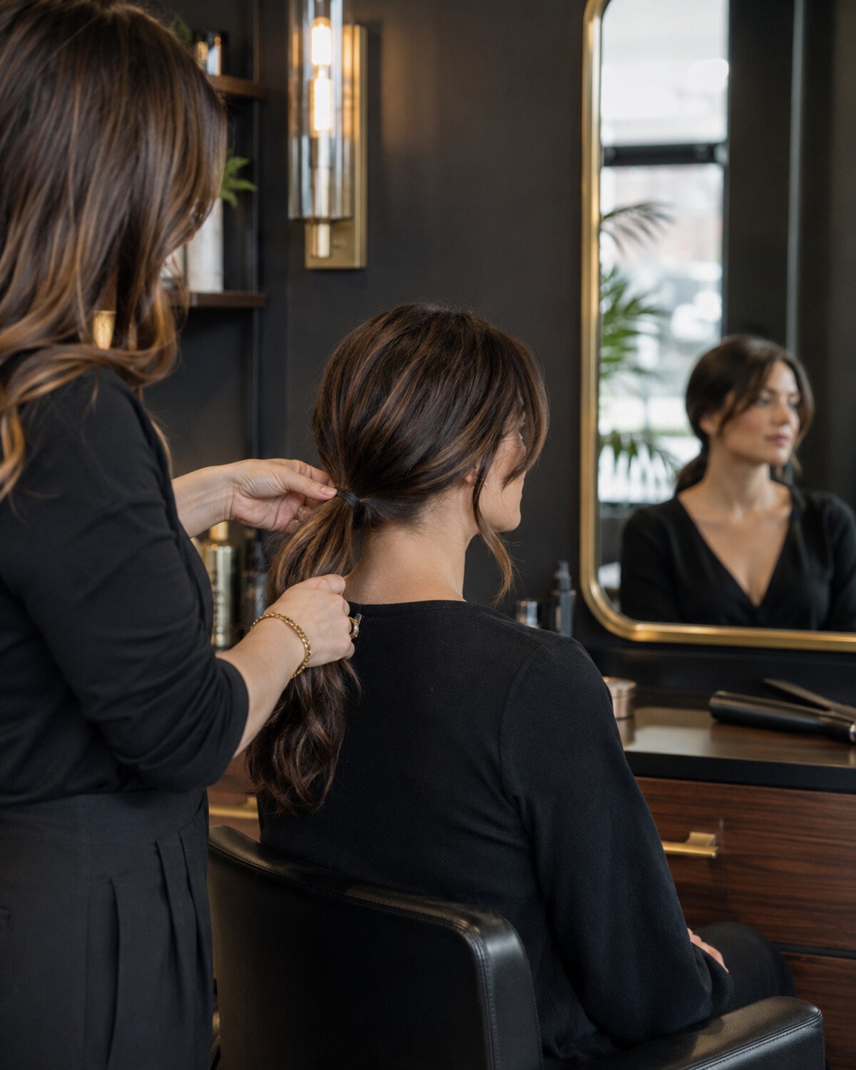 Professional stylist gently gathering a client’s long brunette hair into a low ponytail